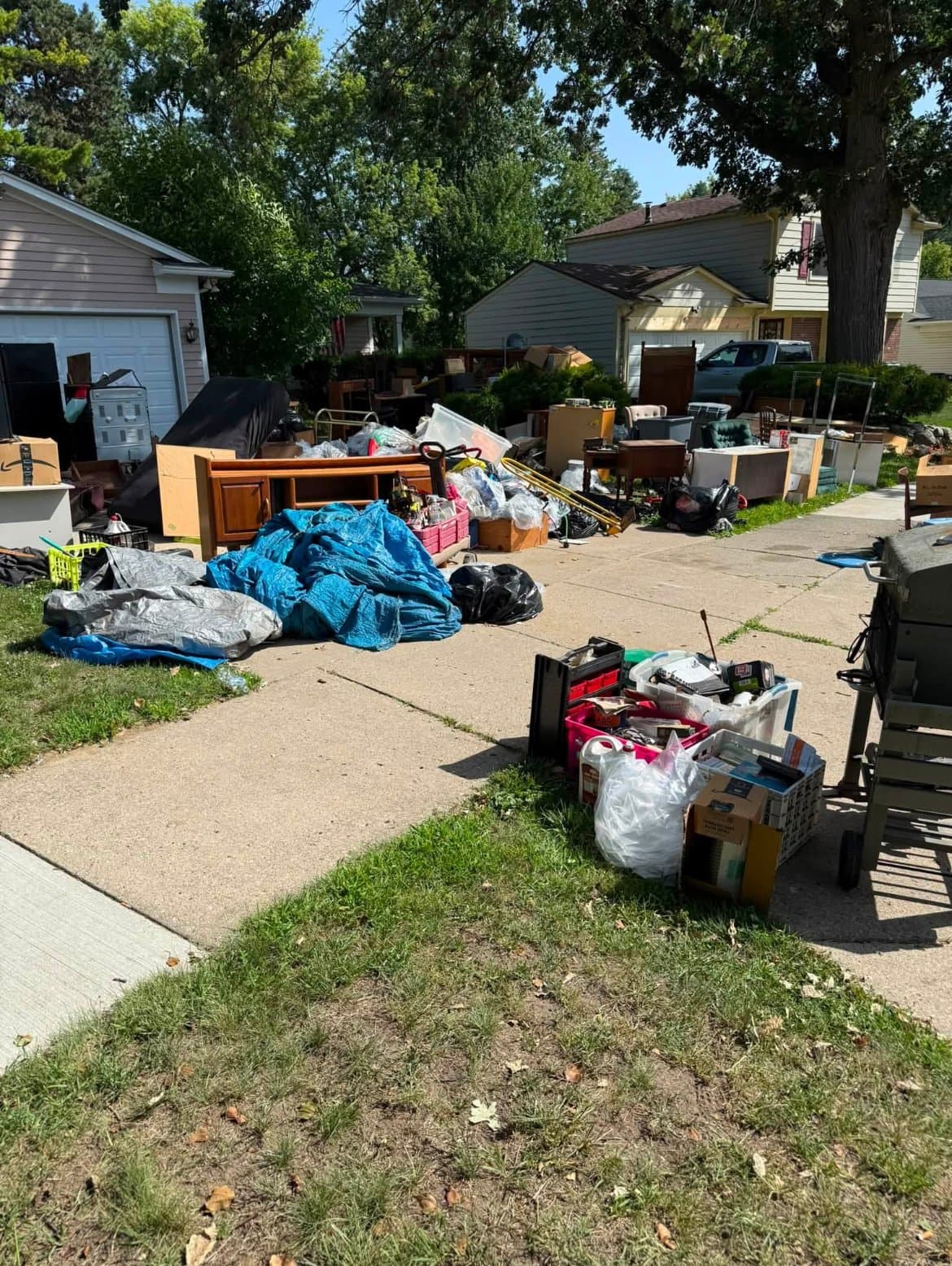 Before: Furniture, tarps, boxes, and household contents piled across a driveway and front lawn in front of a beige garage.