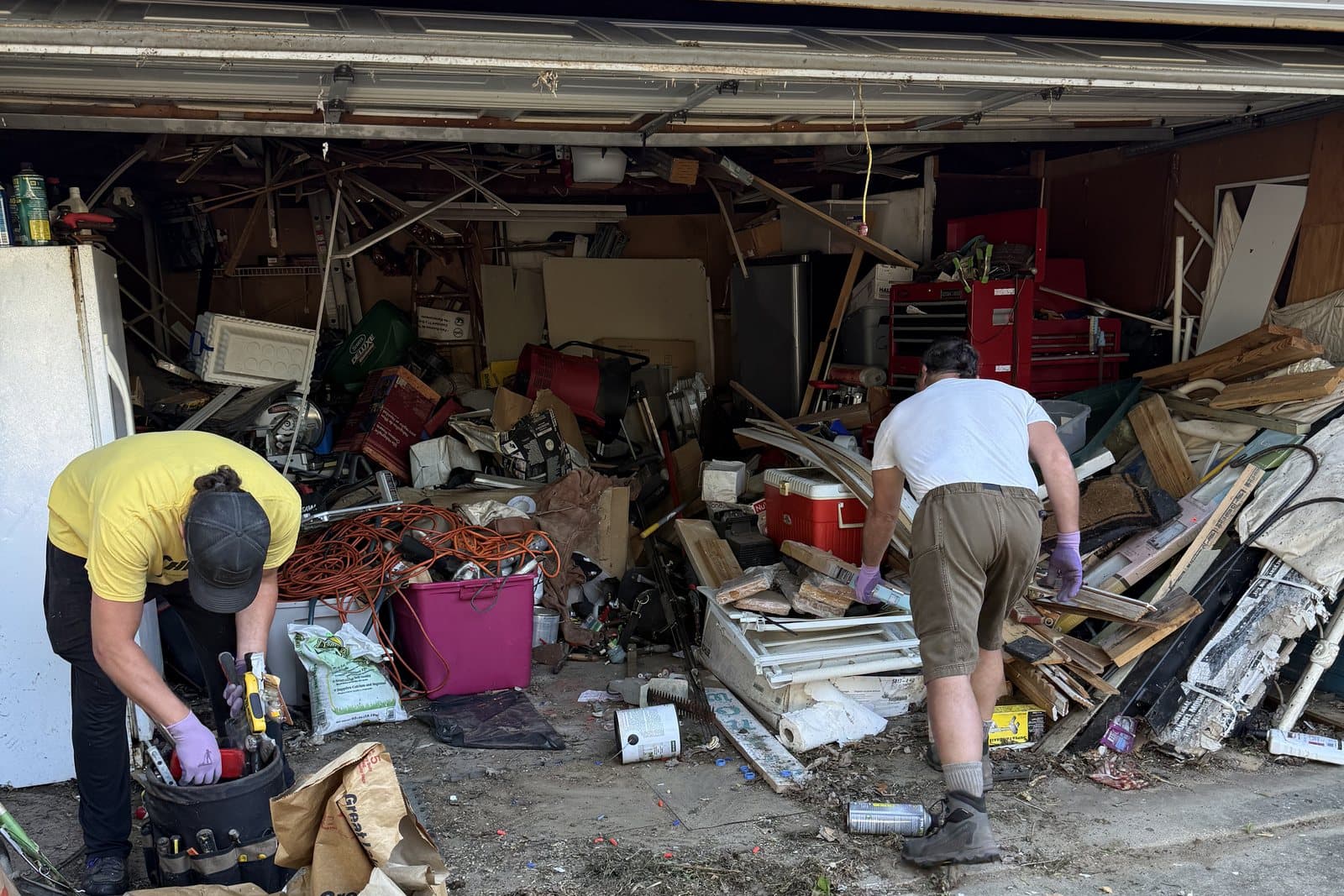 Before: Two workers sorting through a garage packed to the door line with lumber, bins, tools, and stored goods.