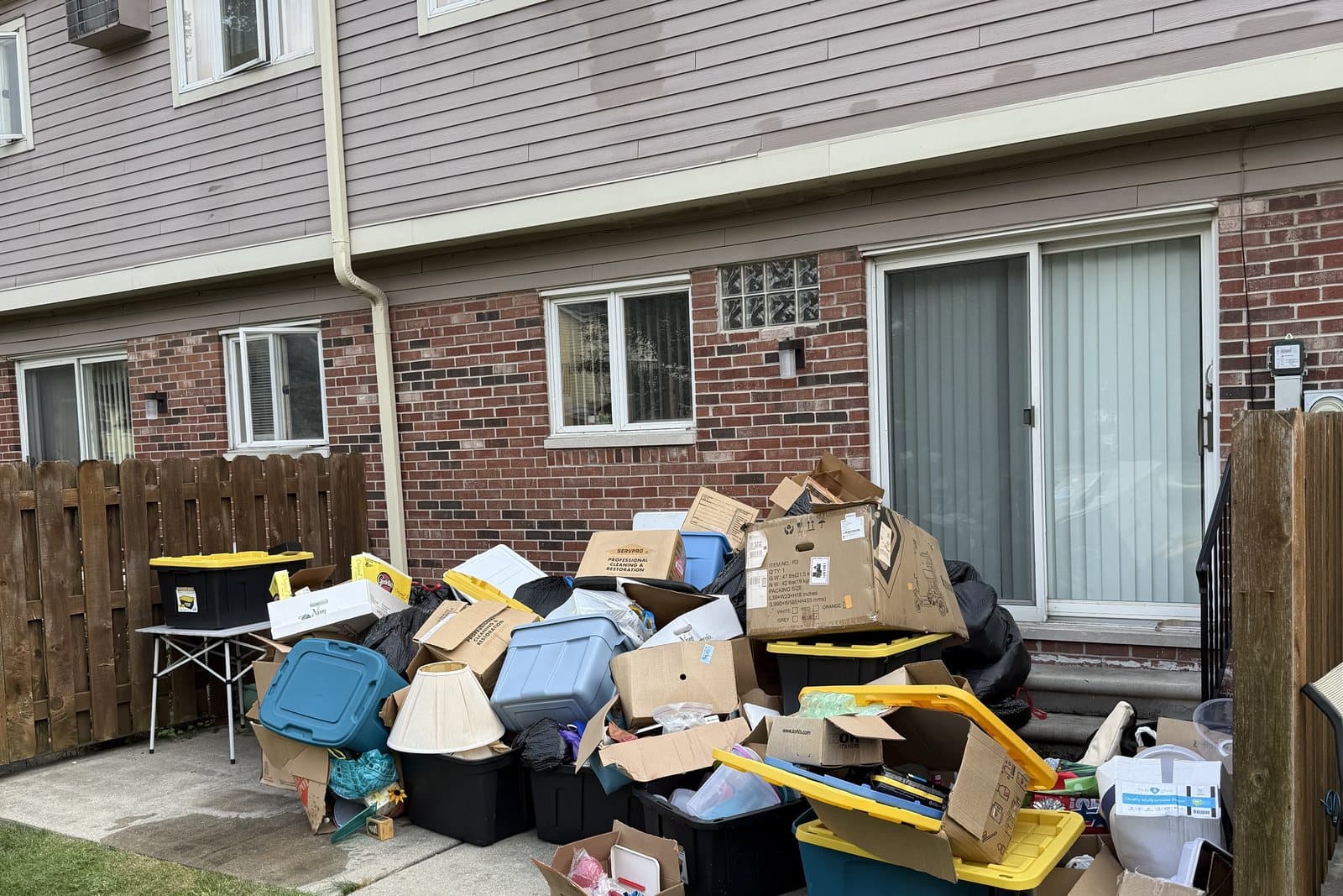 Before: Boxes, storage bins, and household contents piled on a townhouse patio beside a wooden privacy fence.
