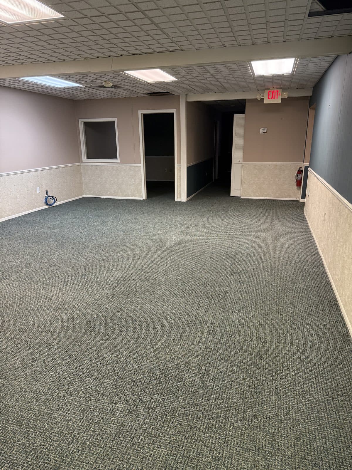 A carpeted open office area with a suspended tile ceiling and a lit exit sign, before demolition.